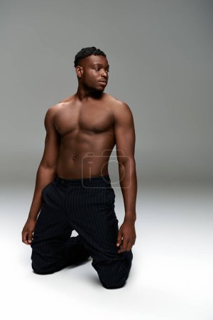 sportive african american man in black pants with strong shirtless body posing on knees on grey
