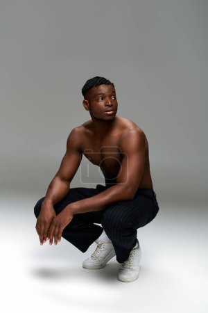 hemdloser muskulöser afrikanisch-amerikanischer Mann in schwarzer Hose sitzt auf den Hinterbeinen und schaut weg auf grau