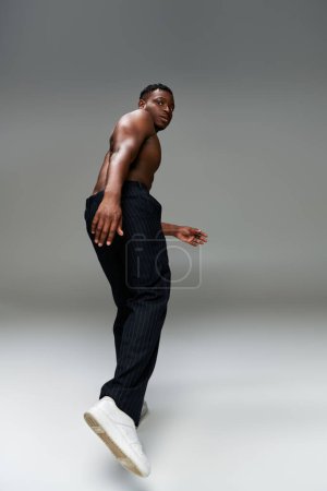 shirtless and strong african american guy in black pants looking at camera while posing on grey