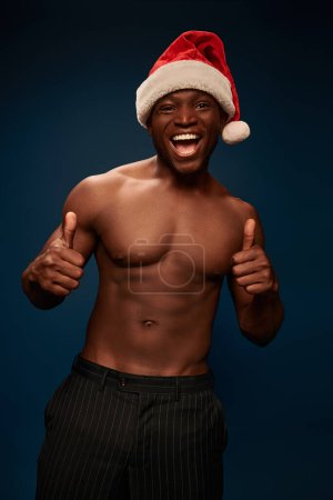 excited and shirtless african american man in santa hat showing thumbs up on navy blue backdrop