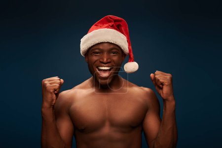 alegre afroamericano hombre con torso muscular con sombrero de santa y regocijo en azul oscuro