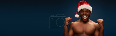 cheerful sportive african american in santa hat showing win gesture on navy blue backdrop, banner