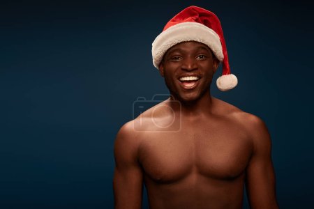glücklich und hemdlos afrikanisch-amerikanischer Kerl mit Weihnachtsmütze lächelt in die Kamera auf marineblauem Hintergrund
