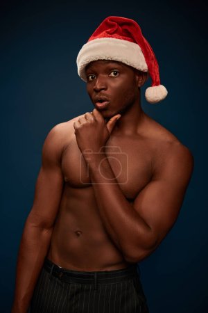 surprised african american man in santa hat with shirtless body looking at camera on dark blue