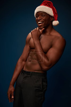 muscular and shirtless african american man in santa hat laughing at camera on navy blue backdrop