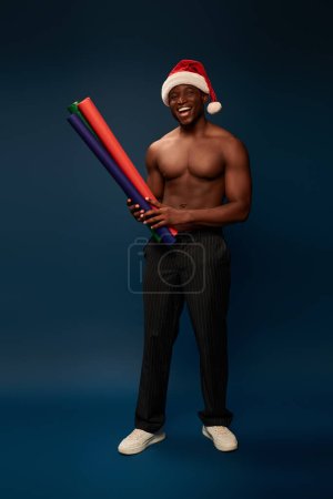 fröhliche muskulöse afrikanisch-amerikanische Mann mit Weihnachtsmütze mit bunten Tapeten auf marineblauem Hintergrund