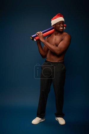 homme afro-américain joyeux et sportif en bonnet de Père Noël avec des rouleaux de papier colorés en studio bleu foncé