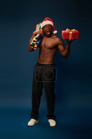 hemdlos und erstaunt afrikanisch-amerikanischer Mann in Weihnachtsmütze mit Tapeten und Geschenk auf dunkelblau
