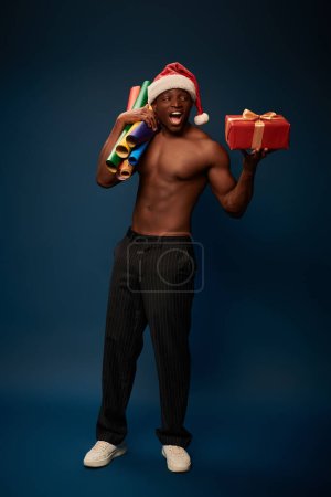 homme afro-américain excité et sportif en chapeau de Père Noël avec boîte cadeau et rouleaux de papier sur bleu foncé