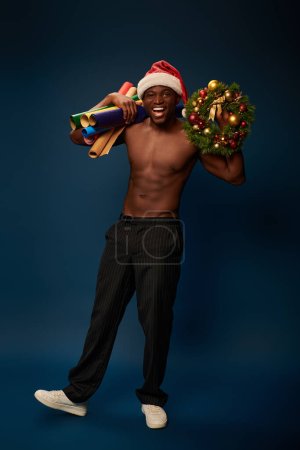 sportive african american man in santa hat with wallpapers and christmas wreath smiling on navy blue