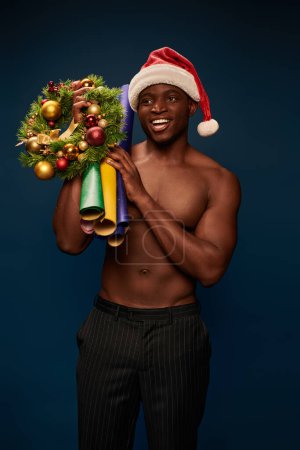 athletische afrikanisch-amerikanische Mann mit Weihnachtsmütze mit Tapeten und Weihnachtskranz lächelnd auf dunkelblau
