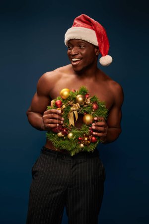 positive shirtless african american man in santa hat with christmas wreath looking away on dark blue