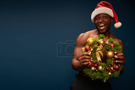 amazed athletic african american man in santa hat with christmas wreath looking at camera in studio