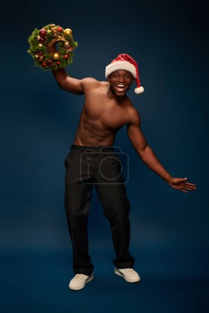 Foto de Alegre y sin camisa hombre afroamericano en sombrero de santa posando corona de Navidad en azul marino - Imagen libre de derechos