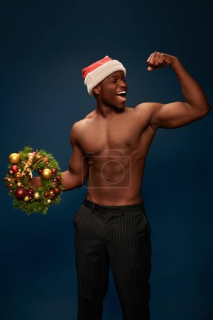 strong african american man in santa hat with christmas wreath demonstrating strength on dark blue