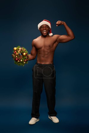astonished and athletic african american man in santa hat showing christmas wreaths on navy blue