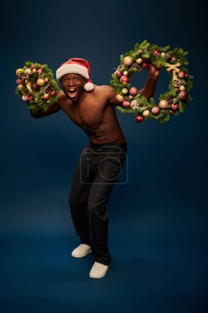 impressed and muscular african american man in santa cap holding christmas wreaths on navy blue