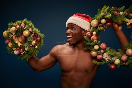 aufgeregtes hemdloses afrikanisch-amerikanisches Männchen in Santa Hap steht mit Weihnachtskränzen auf dunkelblau