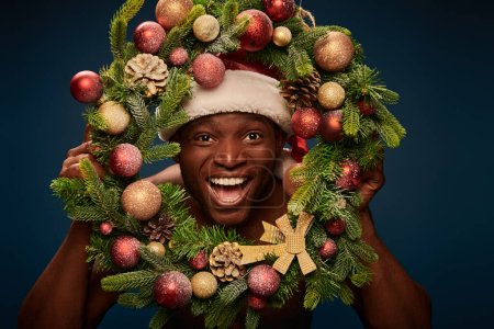 overjoyed african american man in santa hat looking at camera through christmas wreath on dark blue