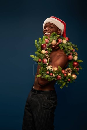 modelo afroamericano caliente y musculoso en sombrero de santa posando con corona de Navidad en azul marino