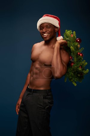 shirtless sportive african american model in santa cap posing with christmas wreath on dark blue