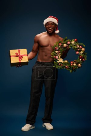 smiley sportive african american man in santa hat holding gift box and christmas wreath on navy blue