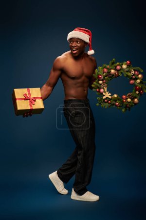 tricky african american man in santa hat walking with present and christmas wreath on navy blue