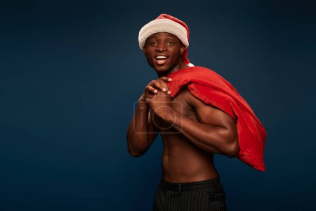 aufgeregter und starker afrikanisch-amerikanischer Mann mit Weihnachtsmütze, der eine Weihnachtstasche in der Hand hält und auf dunkelblau lächelt