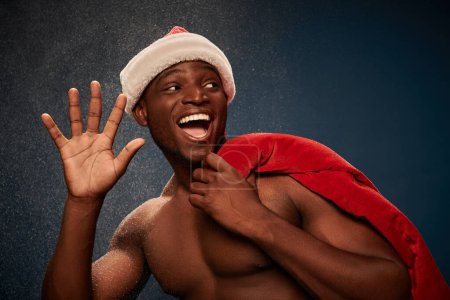 überglücklicher sportlicher afrikanisch-amerikanischer Mann mit Weihnachtsmanntasche, der vor marineblauer, schneebedeckter Kulisse die Hand winkt