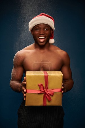 entzückter und muskulöser afrikanisch-amerikanischer Mann mit glänzendem Weihnachtsgeschenk auf dunkelblauem Schneehintergrund