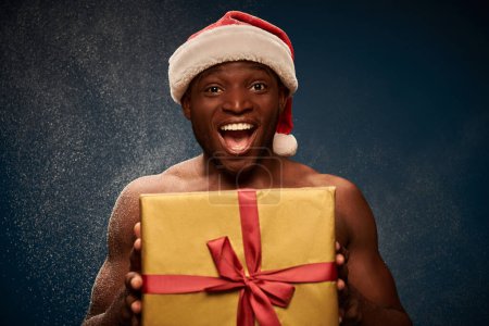 thrilled sportive african american man in santa hat with shiny gift box on dark blue snowy backdrop