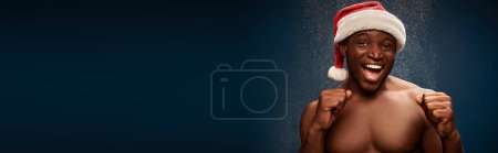 Photo for Joyful athletic african american guy in santa hat showing win gesture on dark snowy backdrop, banner - Royalty Free Image