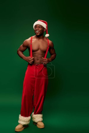 alegre musculoso afroamericano hombre en sombrero de santa y pantalones rojos con tirantes sobre fondo verde