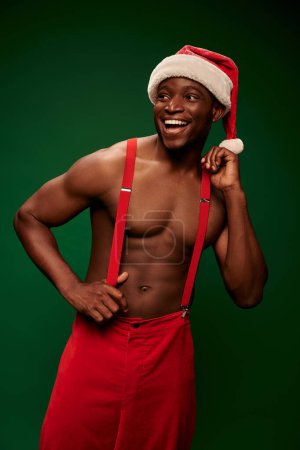joyful shirtless african american man in santa hat and red pants looking away on green backdrop