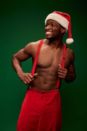 excited shirtless african american guy in santa hat and red pants looking away on green background