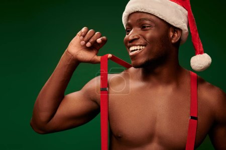 athletischer afrikanisch-amerikanischer Mann mit Weihnachtsmütze und Hosenträgern, der lächelt und wegschaut