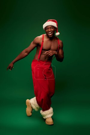 excited athletic african american man in santa hat and red pants on suspenders posing on green