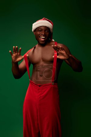 shirtless muscular african american guy in santa hat pants pulling suspenders and smiling on green