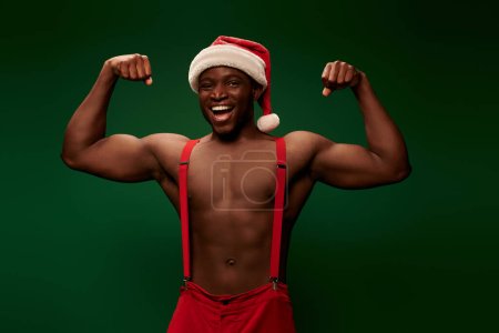 happy and strong african american man in christmas hat and pants showing muscles on green backdrop