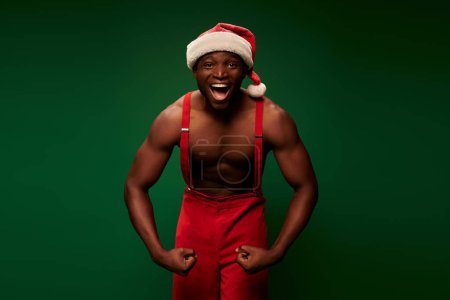 Foto de Hombre afroamericano energizado y sin camisa en sombrero de Navidad y pantalones que muestran músculos en verde - Imagen libre de derechos