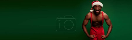 Photo for Athletic african american man in christmas hat and red pants showing muscles on green, banner - Royalty Free Image