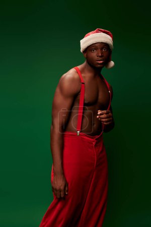 nachdenklicher afrikanisch-amerikanischer Mann mit schulterfreiem muskulösem Oberkörper in Weihnachtsmütze und roter Hose auf grün