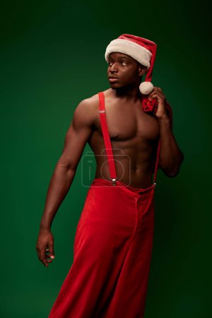 junger, hemdloser afrikanisch-amerikanischer Typ mit Weihnachtsmütze und Hose mit Hosenträgern, die auf grün wegschauen
