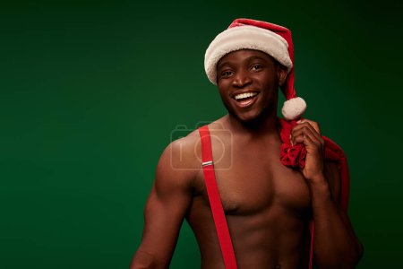 muscular african american man in santa hat holding christmas bags and looking at camera on green