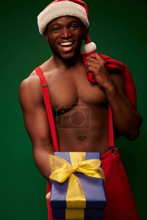 fröhliche hemdlose afrikanisch-amerikanische Mann in Weihnachtsmütze mit Weihnachtstasche zeigt Geschenkbox auf grün