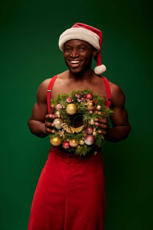 fröhlicher und sportlicher afrikanisch-amerikanischer Mann mit Weihnachtsmütze und roter Hose mit Weihnachtskranz auf grünem Grund