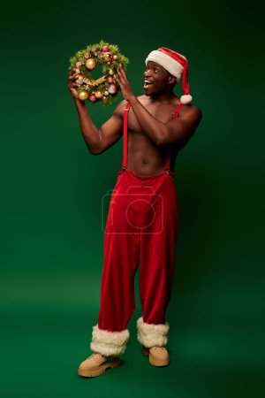 amazed african american man in santa hat and red pants looking at shiny christmas wreath on green