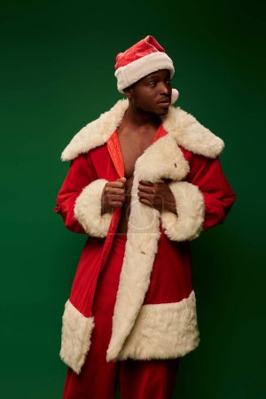 handsome african american man in santa claus costume standing and looking away on green backdrop