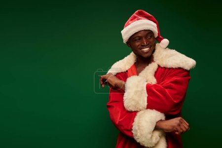 joyful african american guy in festive santa claus costume looking away on green backdrop