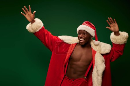 fröhlicher afrikanisch-amerikanischer Kerl im Weihnachtskostüm auf hemdlosem Körper, der Hände vor grünem Hintergrund wedelt